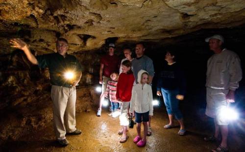 A group of people in a cave, listening to a guide while holding flashlights. Stalactites and rock formations are visible.