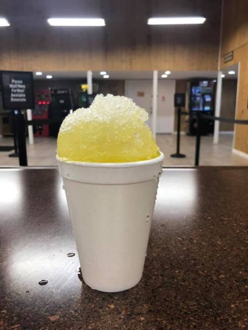 A white foam cup filled with yellow slushy ice, sitting on a brown table in a casual indoor setting.