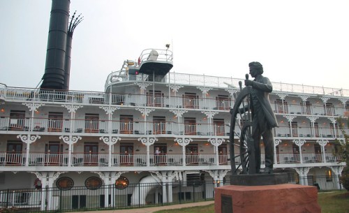 A riverboat with a tall smokestack and a statue of a man at a ship's wheel in the foreground.