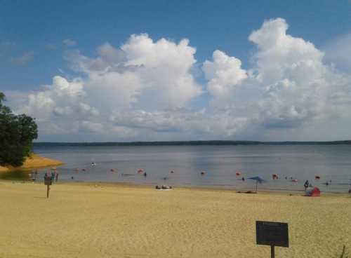 A sandy beach with people swimming and relaxing, under a blue sky filled with fluffy white clouds.