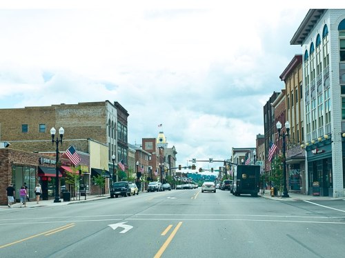 A view of a downtown street lined with shops and flags, featuring a clock tower in the background under a cloudy sky.