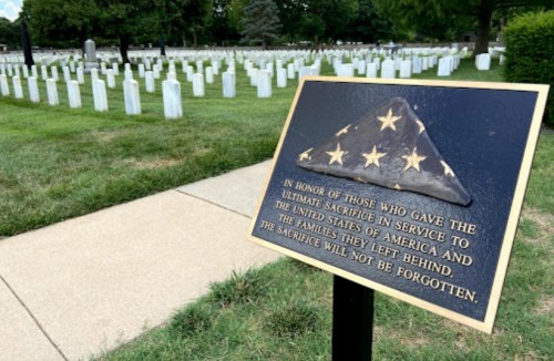 A plaque honoring those who sacrificed in service to the U.S., with a cemetery of white gravestones in the background.