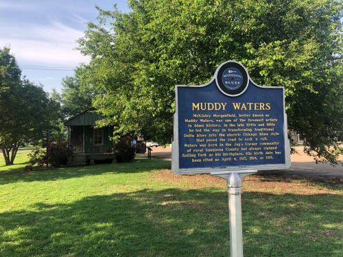 Historical marker for Muddy Waters, highlighting his impact on blues music, with a small building and trees in the background.