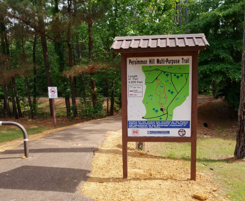 Sign for Persimmon Hill Multi-Purpose Trail, featuring a map and trail information, surrounded by trees.