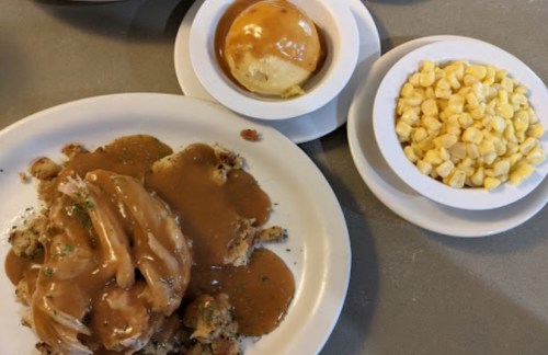 A plate of chicken with gravy over stuffing, served with corn and a roll drizzled with honey.