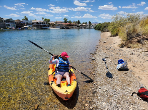 A person in a kayak paddles along a clear river, with a sandy shore and houses in the background under a blue sky.