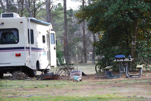 A camper parked in a wooded area with picnic tables and chairs nearby, surrounded by trees and greenery.