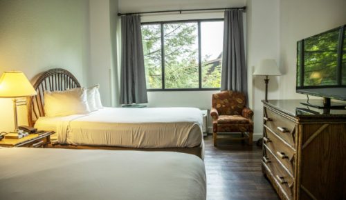 A cozy hotel room with two beds, a window, a chair, and a TV, featuring warm lighting and wooden furniture.