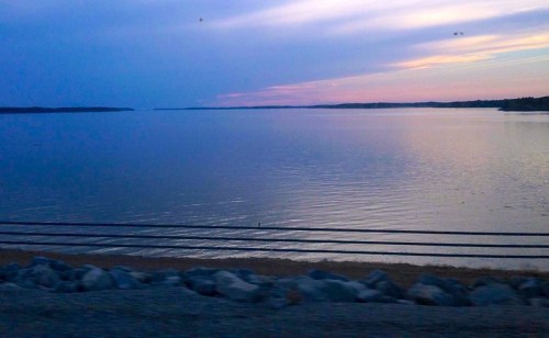 A tranquil lake at dusk, reflecting soft hues of blue and purple, with a rocky shoreline and distant horizon.