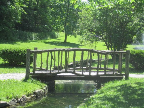 A rustic wooden bridge spans a small stream in a lush green park, surrounded by trees and vibrant grass.