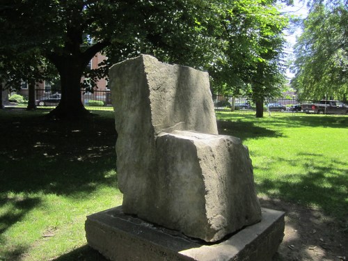 A large, abstract stone sculpture resembling a chair, set in a grassy area under trees.