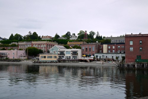 A waterfront view of colorful buildings along a shoreline, reflecting in calm water under a cloudy sky.