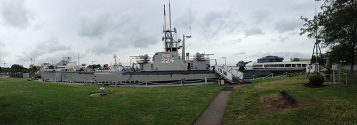 A panoramic view of a large military ship docked near grassy areas and buildings under a cloudy sky.