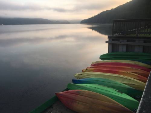 Colorful kayaks lined up on a dock by a calm lake at sunrise, with misty mountains in the background.