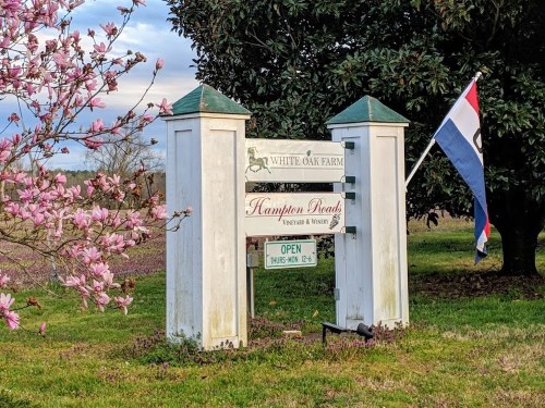 Sign for White Oak Farm and Hampton Roads Vineyard & Winery, with a flag and blooming flowers nearby.