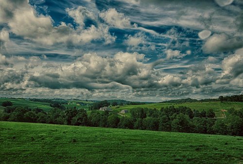 A lush green landscape under a dramatic sky filled with clouds, showcasing rolling hills and distant trees.