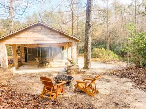 Cozy cabin by a river, featuring a fire pit and wooden chairs, surrounded by trees and autumn leaves.