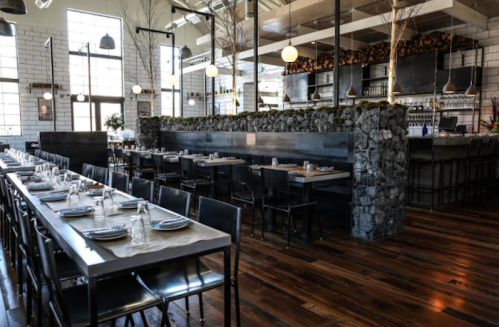 Modern restaurant interior with long tables, metal chairs, and a stone feature wall, featuring warm wood flooring.