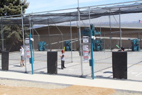 Two children practice baseball swings in a fenced batting cage with pitching machines in the background.