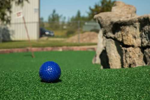 A blue golf ball on artificial green turf, with a rock formation and a distant flag in the background.