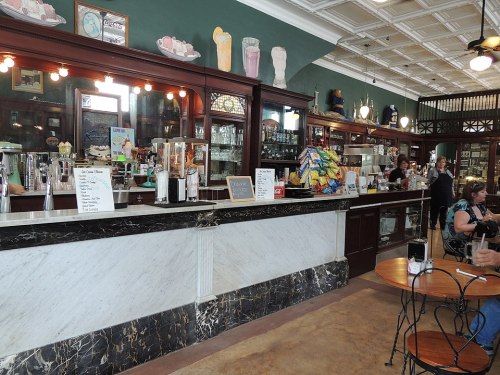 A vintage café interior with a marble counter, shelves of glassware, and patrons enjoying their drinks.