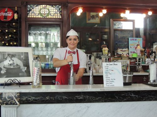 A smiling woman in a retro ice cream shop uniform serves ice cream at a marble counter with vintage decor.