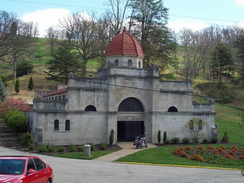 A stone building with a red dome roof, surrounded by greenery and a parking area, featuring decorative architectural details.