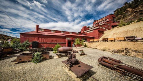 Historic red mining building with equipment and rail cars, set against a cloudy sky and rocky landscape.