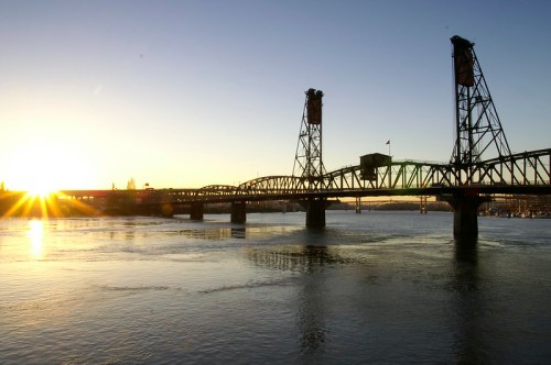 Sunrise over a bridge spanning a river, with sunlight reflecting on the water and a clear sky in the background.