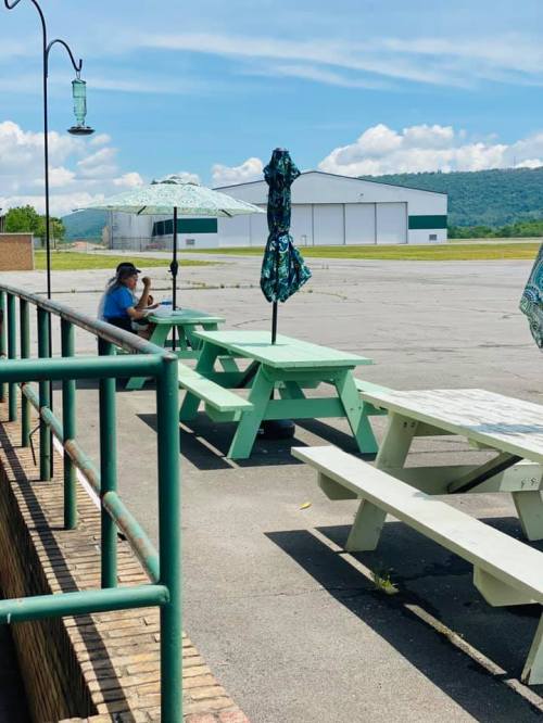 A person sits at a picnic table under an umbrella, with a hangar and mountains in the background on a sunny day.