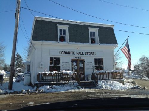 A quaint, snow-covered store with a sign reading "Granite Hall Store," featuring an American flag and holiday decorations.