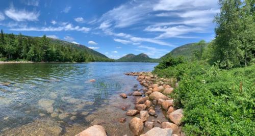 A serene lake surrounded by green trees and rocky shores, with mountains in the background under a blue sky.