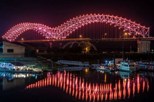 A brightly lit bridge arches over a calm river at night, reflecting colorful lights on the water's surface.