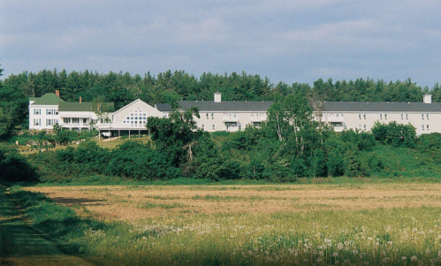 A scenic view of a white building complex surrounded by greenery and fields under a cloudy sky.