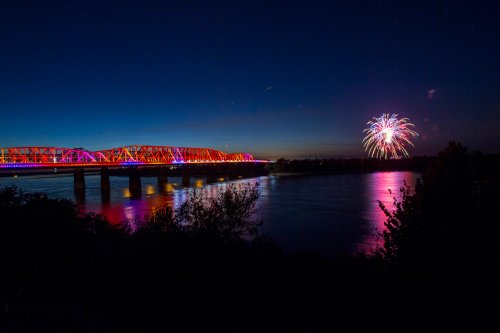 A colorful bridge illuminated at night with fireworks in the sky above a river.
