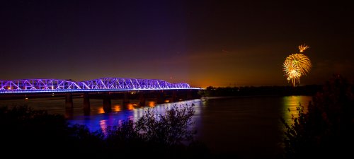 A brightly lit bridge at night with colorful fireworks in the sky, reflecting on the water below.