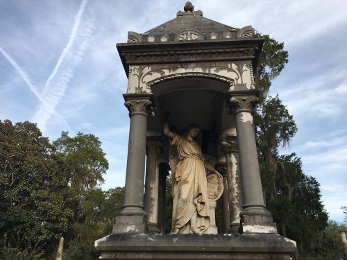 A stone mausoleum with a statue of a figure holding a shield, surrounded by trees and a cloudy sky.