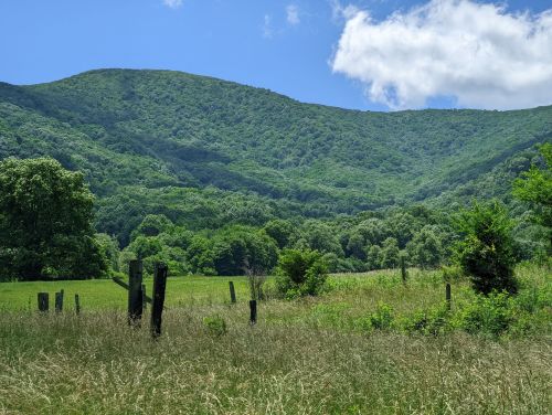 Lush green mountains under a blue sky with scattered clouds, framed by a grassy field and wooden posts in the foreground.
