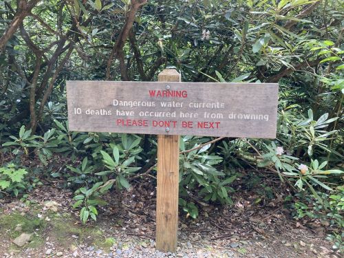 A wooden warning sign about dangerous water currents, noting 10 drowning deaths and urging caution.