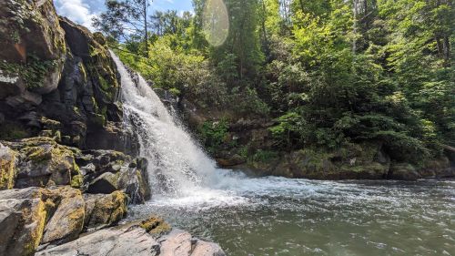 A serene waterfall cascades over rocks into a clear pool, surrounded by lush green trees and a bright blue sky.