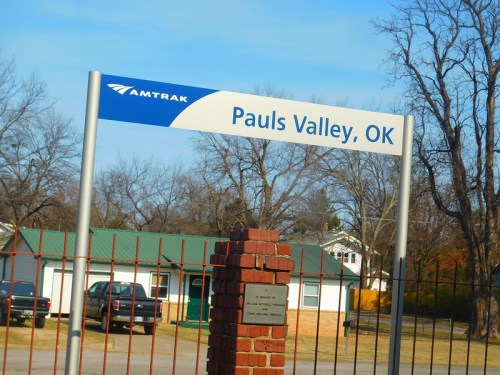 Sign for Amtrak station in Pauls Valley, OK, with a brick base and trees in the background.