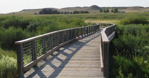 A wooden boardwalk curves through green marshland toward a brown building and grassy hills under a bright sky.