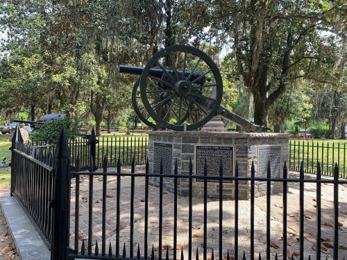 A historic cannon on a stone pedestal, surrounded by a black fence in a park with trees and greenery.