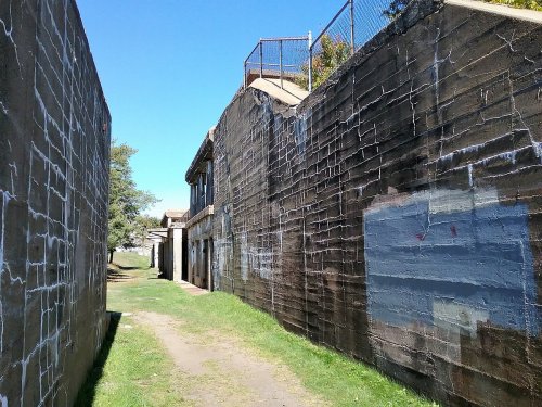 A narrow pathway between two weathered stone walls, leading to a building under a clear blue sky.