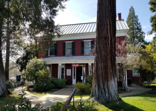 A red two-story house with green shutters, surrounded by trees and greenery, under a clear blue sky.
