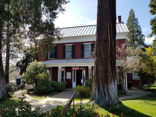 A red two-story house with green shutters, surrounded by trees and greenery, under a clear blue sky.