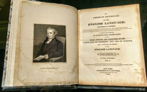 An open book displaying the title page of "An American Dictionary of the English Language" by Noah Webster, with a portrait.