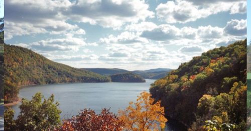 A scenic view of a river surrounded by hills, with colorful autumn foliage and a cloudy blue sky.