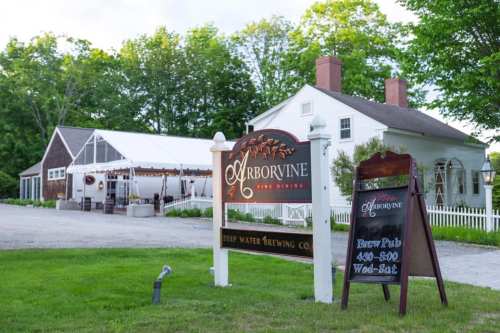 A sign for Arborvine, a beer pub, in front of a white building with a tent and green trees in the background.