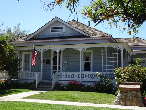 A charming blue house with white trim, a front porch, and an American flag, surrounded by greenery and orange trees.